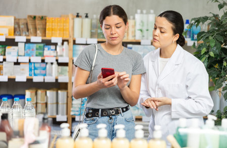 Young girl scrolling phone while looking for something and pharmacist consultingの写真素材