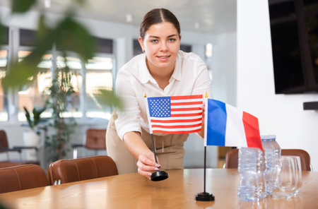 Positive young woman putting flag of the USA on the table with flag of France in conference room