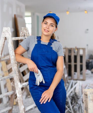 Portrait of contented young female builder in blue overalls during the repair of cottage