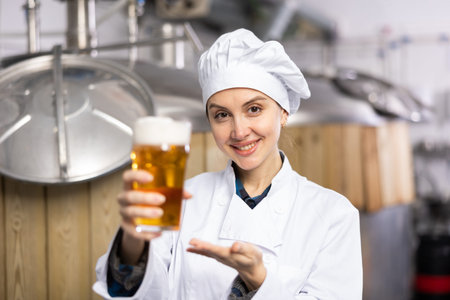 Portrait of female brewer who is standing with glass of beer on her workplace in brew-house