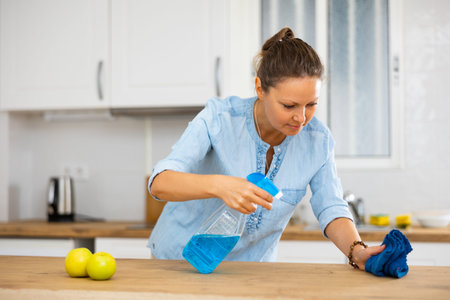 Female in a blue blouse rubbing kitchen table with a rag while using household chemicals