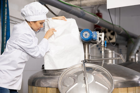 Female brewer pouring malted grain from bag into fermenter in breweryの写真素材