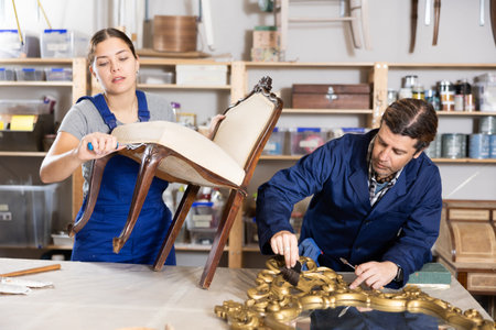 Male restorer treats wooden frame of vintage mirror with protective varnish. While woman restorer...