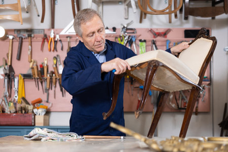 Experienced male restorer removing upholstery of vintage chair for restoration in workshop