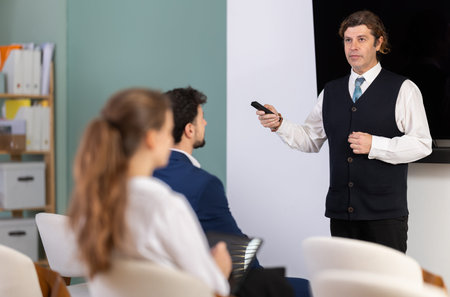 Male lecturer communicates and addressed to attendees, controls projector using remote control