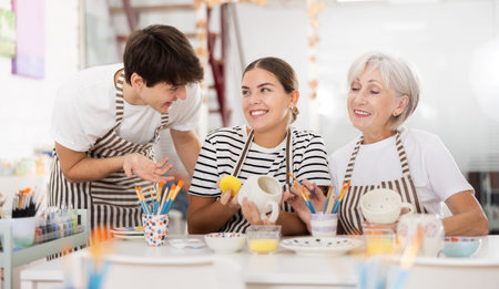 Cheerful older woman with young adult children painting pottery in family workshop