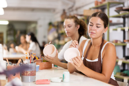Young woman sits near table and decorates clay ceramic craft vase, plate
