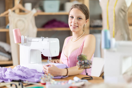 Positive young female dressmaker working with sewing-machine in tailors workshop