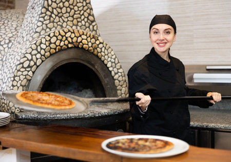 Portrait of female chef in black uniform holding big shovel with pizza in kitchen
