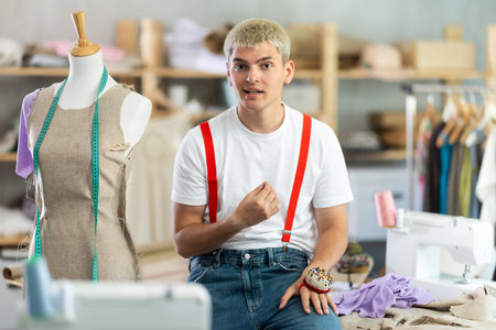 Experienced young guy designer posing near work place with mannequin and sewing machine and tools,...