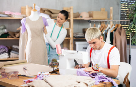 Young guy tailor sewing textile using sewing machine and asian female assistant working with fabric...