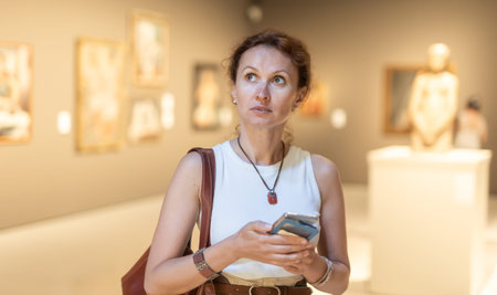 Portrait of woman visitor with smartphone in hands looking exhibition of paintings in museum