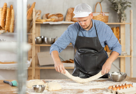 Professional baker prepares raw yeast dough in a bakery, shaping it into baguette