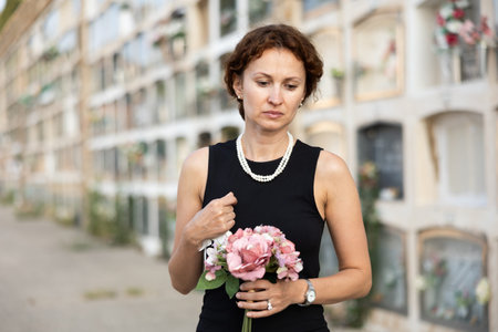 Woman in black dress with bouquet of flowers came to cemetery to honor the memory of deceased loved...