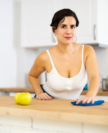 Smiling young Hispanic woman wiping kitchen surfaces at home
