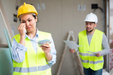 Upset female builder standing in building under renovation with money