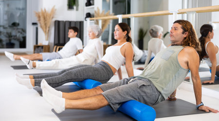 Positive athletic male performing set of exercises with pilates roller during group class in fitness...