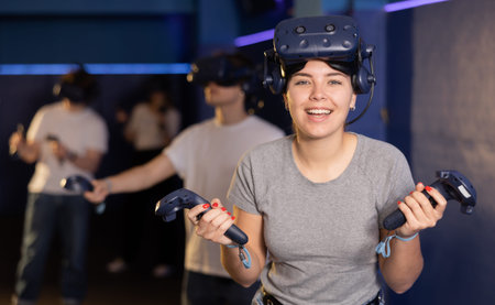 Cheerful young girl with VR headset and joysticks playing with friends