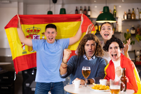 Excited young adult sports fans waving flag of Spain and supporting national team with beer in pubの写真素材