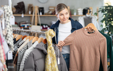 Fashionable european woman in casual clothes standing near clothes rack and choosing new dress for...