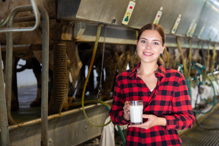 Young female worker standing near milking line with glass of milk