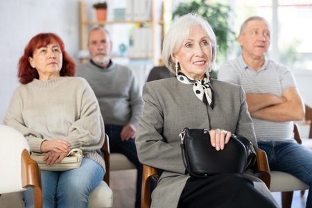 Cheerful and sociable mature woman sitting on chairs, listening and laughing with group of people of...