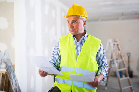 Portrait of worker in a yellow vest with documents in his hands in renovated cottage