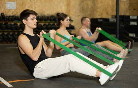 Motivated young man practicing exercises with stretch rope sitting near other people in gym