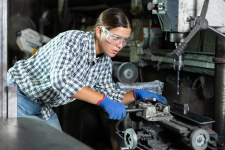 Girl worker at metallurgical plant drills hole in metal plate using drilling machine