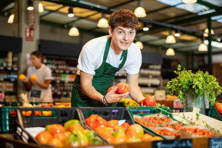 Cheerful guy seller arranging ripe tomatoes on counter in grocery store
