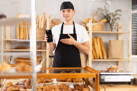 Portrait of friendly male baker with smartphone in hands in bakery interior