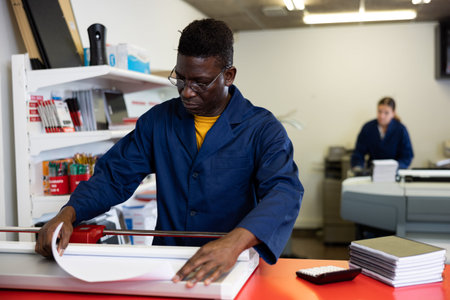 Focused man in blue uniform trimming the printed work on the cutter in the typography