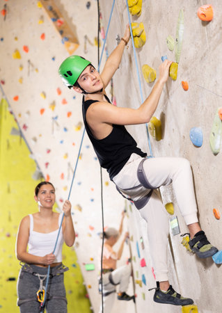 young positive man, insured by trainer, moving up artificial climbing wall indoors