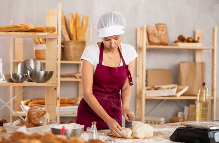 Focused young female baker in apron working at bakery, cutting dough into pieces