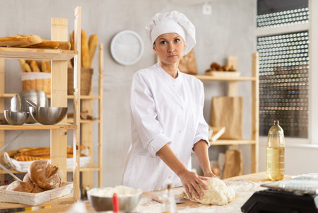 Positive young female baker in white uniform kneading dough and preparing dough for baking at...