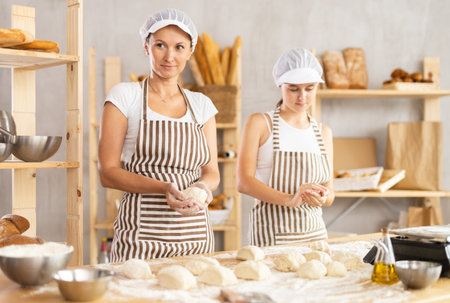 Female baker and her young assistant roll dough into balls to make delicious buns