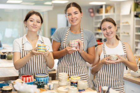 Three positive girls stands in pottery workshop and show created work, finished decorative vase