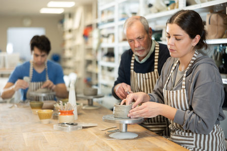 Elderly man teacher shows girl how to sculpt ceramic product from raw clay in workshop