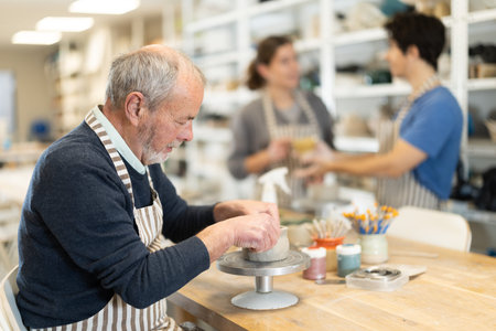 Mature man at table molds cup from wet clay in pottery workshopの写真素材