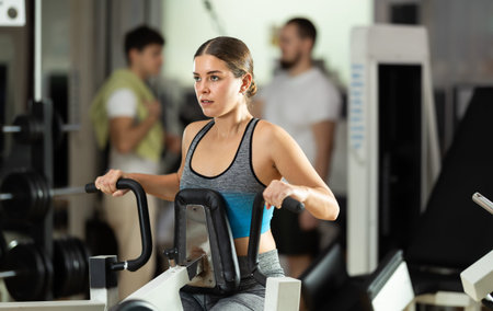Girl in gym performs exercises for arm muscles using simulators and fitness center equipment.