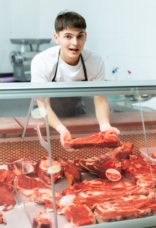Young butcher shop seller arranging fresh beef in glass refrigerated display
