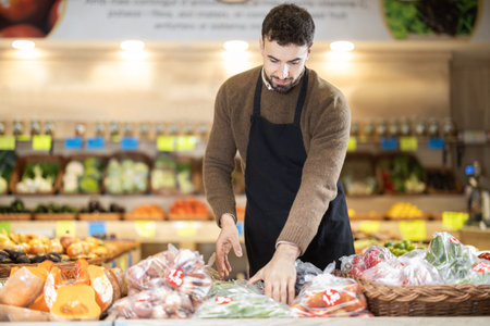 Young male seller putting long green peppers on counter in grocery market