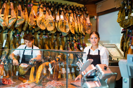 Young female and male vendors arranging jamon at counter in butcher shop