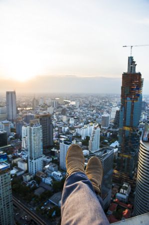 Legs hanging down from the roof top into the air with front aerial view over beautiful twilight of a big city in Bangkok, extreme activity, dangerous, relax.の写真素材