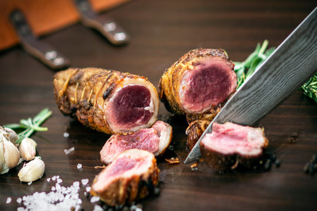 slicing a serving of organic roast beef roll with knife on wood table with garlic pepper and salt in...