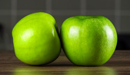 Two shiny green apples are positioned next to each other on a wooden surface