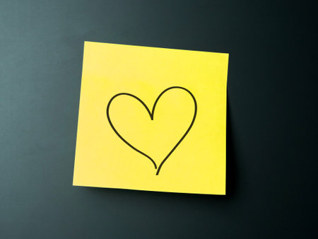 A yellow sticky note features a simple heart drawn in black on a dark surfaceの写真素材