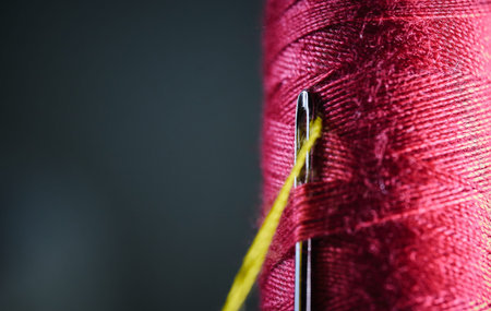 Macro shot of a metal needle eye with yellow thread against a red textile thread spool
