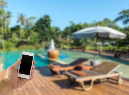 Close up of Male Hand Holding Smart Mobile Phone with Blank Screen on The Blur Swimming Pool with Various Tree in Luxury Hotel used as Template to input Text or Photo, Business Conceptの写真素材