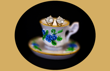 A pair of white diamond stud earrings set in yellow gold, sitting in a miniature teacup on a black oval and yellow backgroundの写真素材